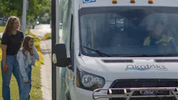 a woman and girl stand on a curb next to an Okotoks Transit cutaway bus a woman and girl stand on a curb next to an Okotoks Transit cutaway bus