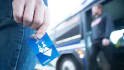 The image shows someone holding a clipper fare card with a person standing in the background blurred out due to focal length. The image shows someone holding a clipper fare card with a person standing in the background blurred out due to focal length.