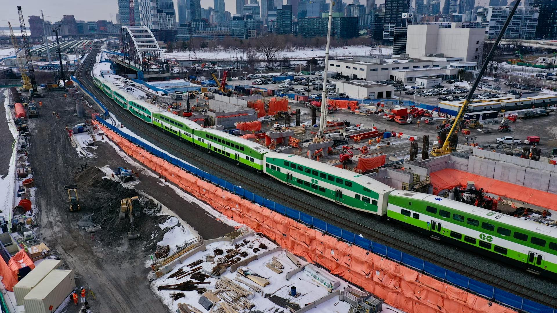 An aerial view of a GO Transit bi-level commuter rail train passing through an active construction zone in winter. The green and white train travels along a corridor flanked by orange safety fencing, construction equipment, cranes, and building materials. The Toronto skyline, including the CN Tower, is visible in the background.