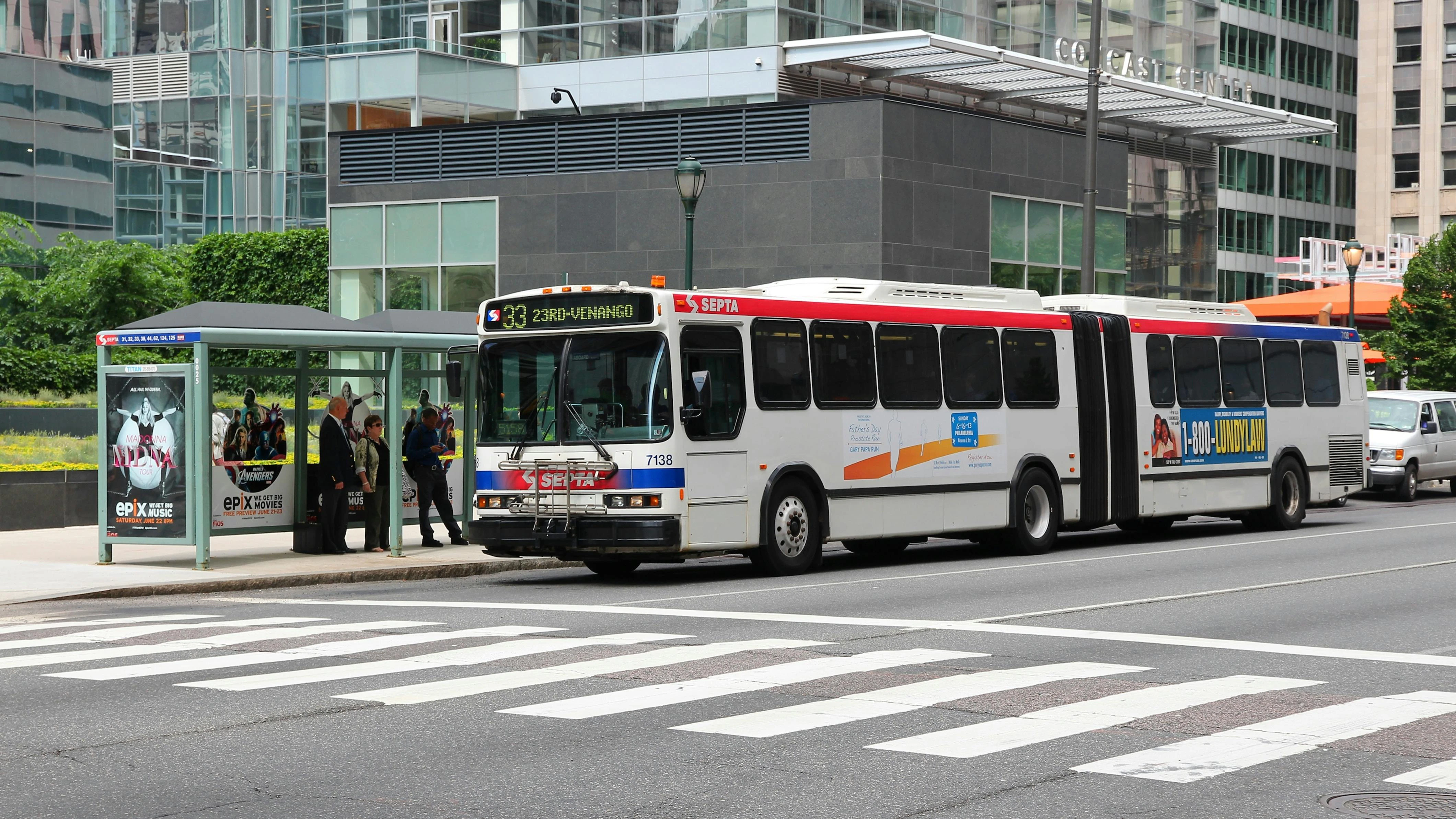 A Southeastern Pennsylvania Transportation Authority articulated bus, Route 33 to 23rd-Venango, stops at a covered bus shelter in downtown Philadelphia as passengers board. The white bus with red and blue striping carries vehicle number 7138. The Comcast Center tower is visible in the background.