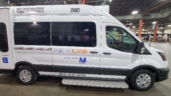 A New Jersey Transit MicroLink van, vehicle number 25801, photographed inside a maintenance facility. The white Ford Transit-based cutaway van displays the MicroLink branding and NJ Transit logo on its side, along with an accessibility symbol indicating wheelchair accessibility. A fold-out entry step is visible below the side door.
