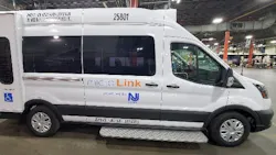 A New Jersey Transit MicroLink van, vehicle number 25801, photographed inside a maintenance facility. The white Ford Transit-based cutaway van displays the MicroLink branding and NJ Transit logo on its side, along with an accessibility symbol indicating wheelchair accessibility. A fold-out entry step is visible below the side door. A New Jersey Transit MicroLink van, vehicle number 25801, photographed inside a maintenance facility. The white Ford Transit-based cutaway van displays the MicroLink branding and NJ Transit logo on its side, along with an accessibility symbol indicating wheelchair accessibility. A fold-out entry step is visible below the side door.