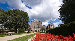 The Ontario Legislative Assembly building at Queen's Park in Toronto, viewed from the front grounds on a sunny summer day. The Romanesque Revival sandstone building is framed by mature trees and a foreground of red and white flowering gardens. A Canadian flag flies from a flagpole to the right of the building. The Ontario Legislative Assembly building at Queen's Park in Toronto, viewed from the front grounds on a sunny summer day. The Romanesque Revival sandstone building is framed by mature trees and a foreground of red and white flowering gardens. A Canadian flag flies from a flagpole to the right of the building.