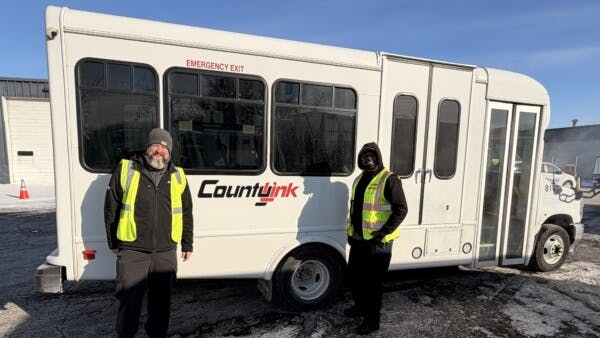 Two transit workers wearing yellow high-visibility vests and winter jackets stand on either side of a white small-transit bus branded with the CountyLink logo. Light snow covers the ground, and a garage or depot building is visible in the background, suggesting a cold-weather operating environment.