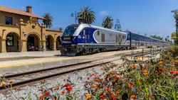 Pacific Surfliner train No. 2112 arrives at Santa Barbara station on a clear day. The blue-and-silver Siemens-built locomotive leads a consist of bilevel passenger cars along the platform of the station's Spanish Colonial Revival depot, its tan stucco walls and red tile roof visible at left. Red and orange lantana blooms fill the foreground, with palm trees and a deep blue sky in the background. Pacific Surfliner train No. 2112 arrives at Santa Barbara station on a clear day. The blue-and-silver Siemens-built locomotive leads a consist of bilevel passenger cars along the platform of the station's Spanish Colonial Revival depot, its tan stucco walls and red tile roof visible at left. Red and orange lantana blooms fill the foreground, with palm trees and a deep blue sky in the background.