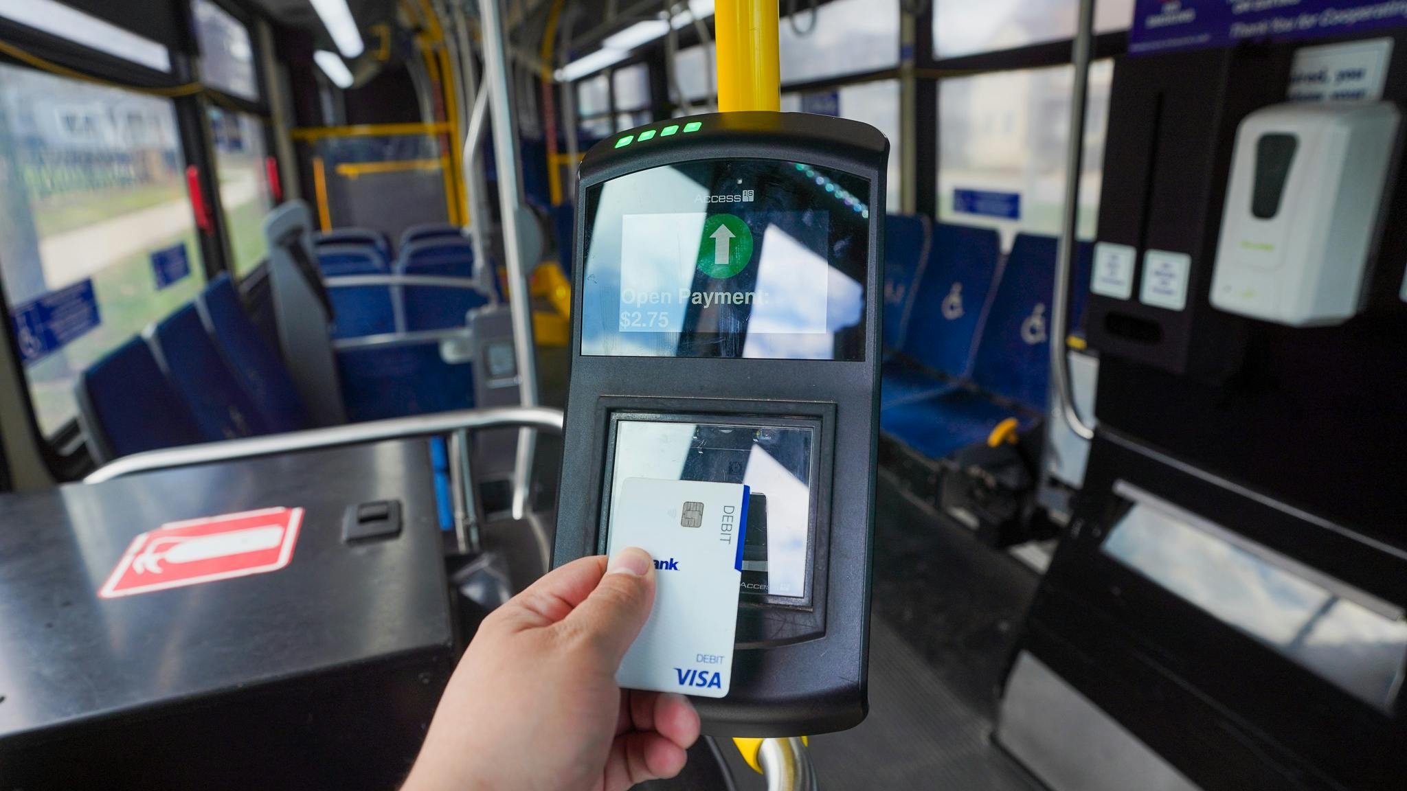 A rider taps a Visa card against a fare reader aboard a Milwaukee County Transit System bus. The reader's screen displays 'Open Payment &ndash; $2.75' with a green upward arrow, indicating a successful contactless transaction. The bus interior, with blue seating and yellow handrails, is visible in the background.