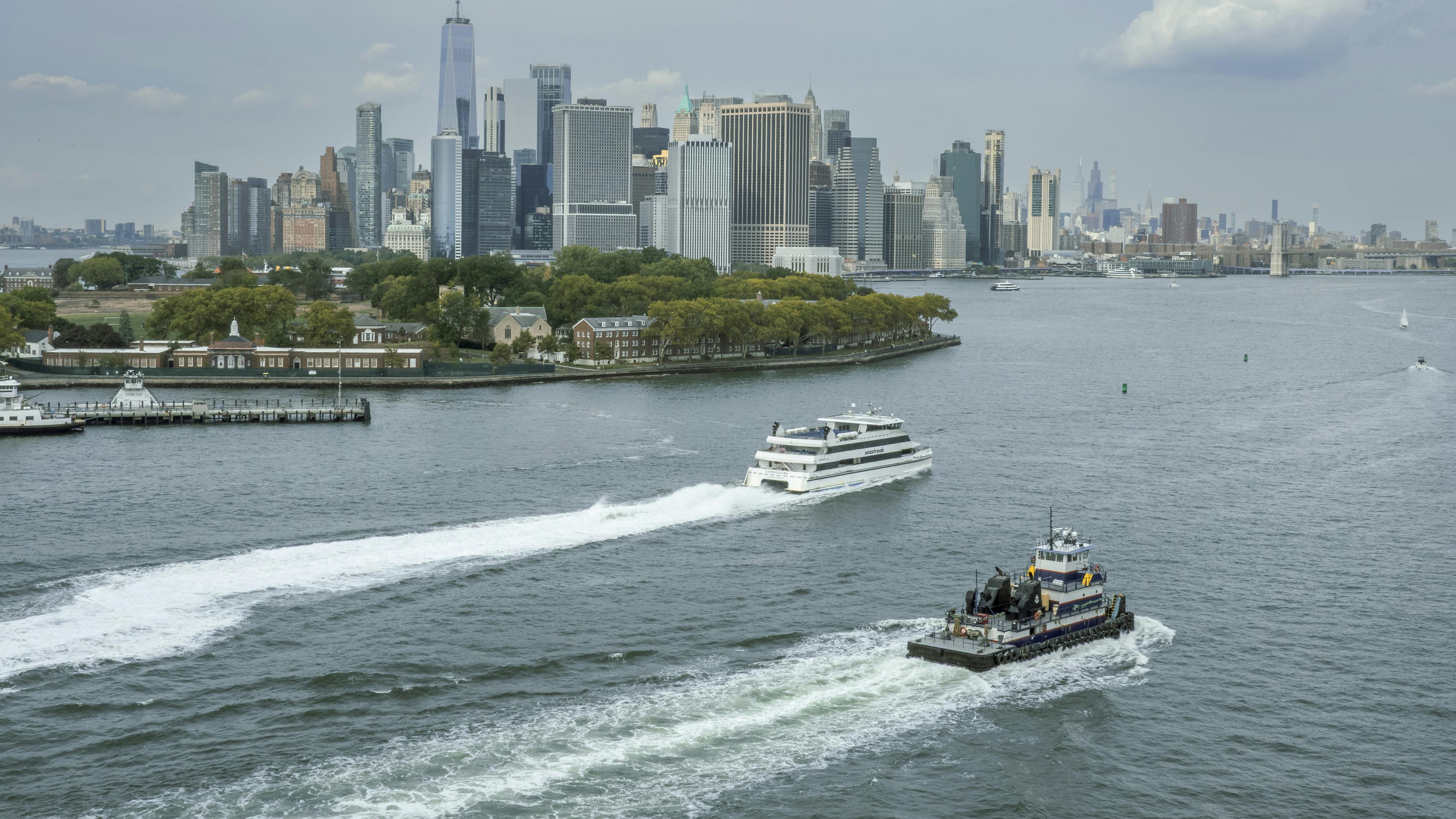Two vessels cross New York Harbor with lower Manhattan's skyline and Governors Island in the background. A white passenger ferry cuts a wake in the middle distance while a smaller tugboat or work vessel passes in the foreground. One World Trade Center rises above the dense cluster of skyscrapers under a partly cloudy sky.