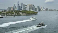 Two vessels cross New York Harbor with lower Manhattan's skyline and Governors Island in the background. A white passenger ferry cuts a wake in the middle distance while a smaller tugboat or work vessel passes in the foreground. One World Trade Center rises above the dense cluster of skyscrapers under a partly cloudy sky. Two vessels cross New York Harbor with lower Manhattan's skyline and Governors Island in the background. A white passenger ferry cuts a wake in the middle distance while a smaller tugboat or work vessel passes in the foreground. One World Trade Center rises above the dense cluster of skyscrapers under a partly cloudy sky.