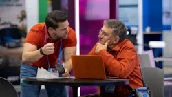 Two conference attendees confer at a small round table on a trade show floor. One person leans in while gesturing, the other listens thoughtfully with a hand to their chin, a laptop open between them. Colorful exhibit booths are visible in the background. Two conference attendees confer at a small round table on a trade show floor. One person leans in while gesturing, the other listens thoughtfully with a hand to their chin, a laptop open between them. Colorful exhibit booths are visible in the background.