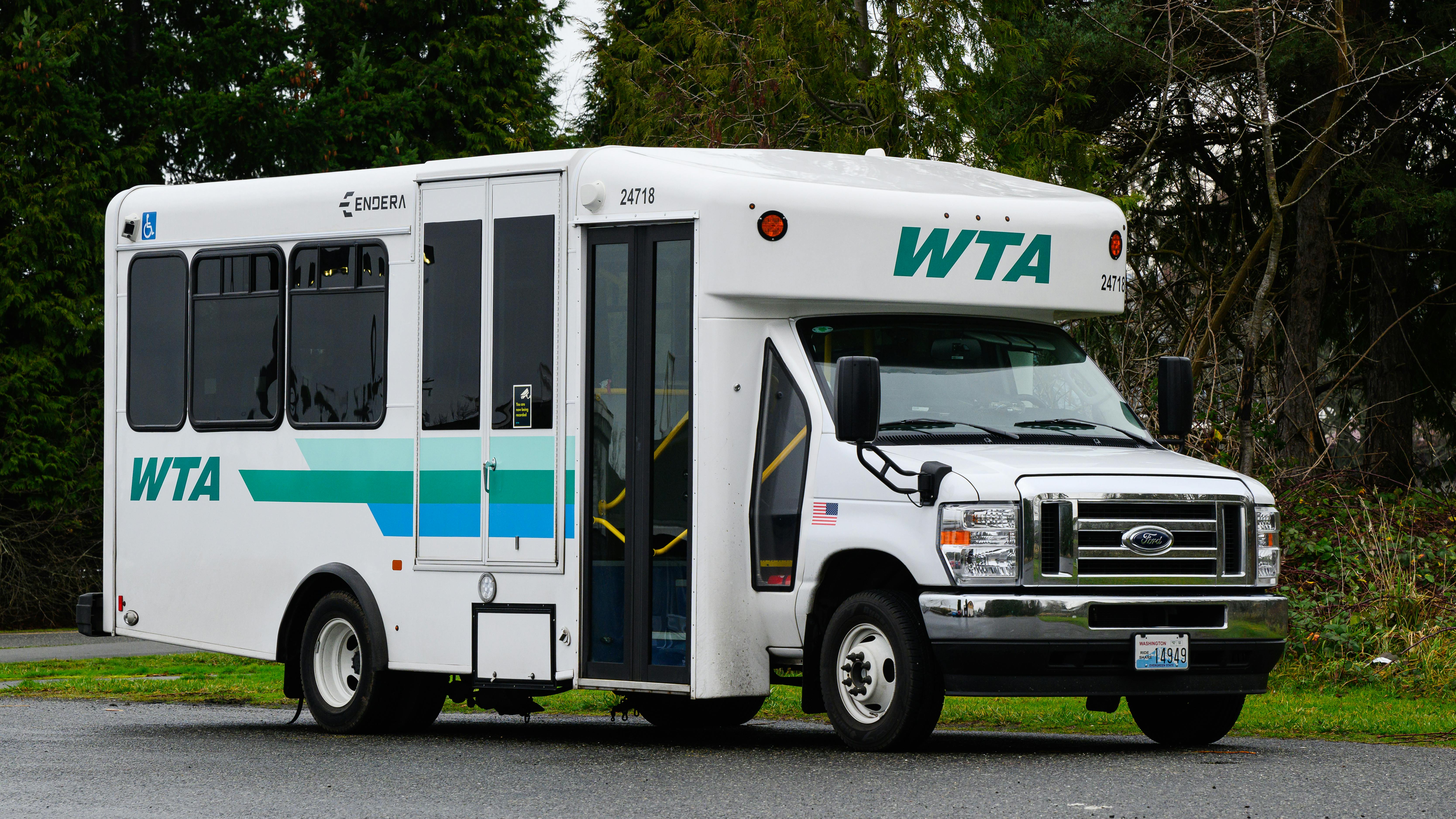 A white WTA (Whatcom Transportation Authority) paratransit/cutaway bus, fleet number 24718, built on a Ford E-Series chassis with an Endera body, photographed in a three-quarter view in a parking area surrounded by evergreen trees. The vehicle features the WTA logo and a teal, green, and blue horizontal stripe livery, a wheelchair accessibility symbol near the front passenger door, and a Washington state license plate. The door is open, revealing yellow interior handrails.