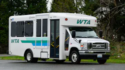 A white WTA (Whatcom Transportation Authority) paratransit/cutaway bus, fleet number 24718, built on a Ford E-Series chassis with an Endera body, photographed in a three-quarter view in a parking area surrounded by evergreen trees. The vehicle features the WTA logo and a teal, green, and blue horizontal stripe livery, a wheelchair accessibility symbol near the front passenger door, and a Washington state license plate. The door is open, revealing yellow interior handrails. A white WTA (Whatcom Transportation Authority) paratransit/cutaway bus, fleet number 24718, built on a Ford E-Series chassis with an Endera body, photographed in a three-quarter view in a parking area surrounded by evergreen trees. The vehicle features the WTA logo and a teal, green, and blue horizontal stripe livery, a wheelchair accessibility symbol near the front passenger door, and a Washington state license plate. The door is open, revealing yellow interior handrails.