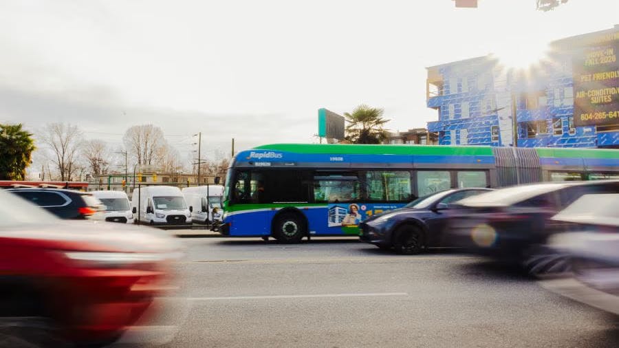 A TransLink RapidBus travels through a busy urban intersection in Metro Vancouver, surrounded by moving vehicles. The bus features the agency's signature green, blue and white livery. Construction of a new apartment building is visible in the background.