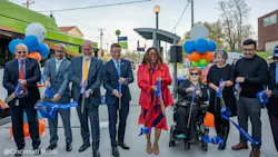 A group of approximately eight officials and community members participate in a ribbon-cutting ceremony at a Cincinnati Metro bus stop, holding blue ceremonial scissors and a blue ribbon reading 'Your Way To Go.' The group stands in front of the Clifton Avenue and Clifton Court stop sign and a green Cincinnati Metro bus. Blue, orange and green balloon clusters flank the group. One participant uses a power wheelchair. The photo is credited to Cincinnati Metro. A group of approximately eight officials and community members participate in a ribbon-cutting ceremony at a Cincinnati Metro bus stop, holding blue ceremonial scissors and a blue ribbon reading 'Your Way To Go.' The group stands in front of the Clifton Avenue and Clifton Court stop sign and a green Cincinnati Metro bus. Blue, orange and green balloon clusters flank the group. One participant uses a power wheelchair. The photo is credited to Cincinnati Metro.