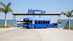 A Big Blue Bus battery-electric bus displaying 'Zero Emissions' on its destination sign sits parked in front of the agency's maintenance and operations facility at 1620 Sixth Street in Santa Monica, Calif., beneath a large 'big blue bus' sign mounted on a canopy at the facility entrance. Additional blue buses are visible parked inside the yard. Palm trees flank the entrance on both sides under a clear blue sky. A Big Blue Bus battery-electric bus displaying 'Zero Emissions' on its destination sign sits parked in front of the agency's maintenance and operations facility at 1620 Sixth Street in Santa Monica, Calif., beneath a large 'big blue bus' sign mounted on a canopy at the facility entrance. Additional blue buses are visible parked inside the yard. Palm trees flank the entrance on both sides under a clear blue sky.