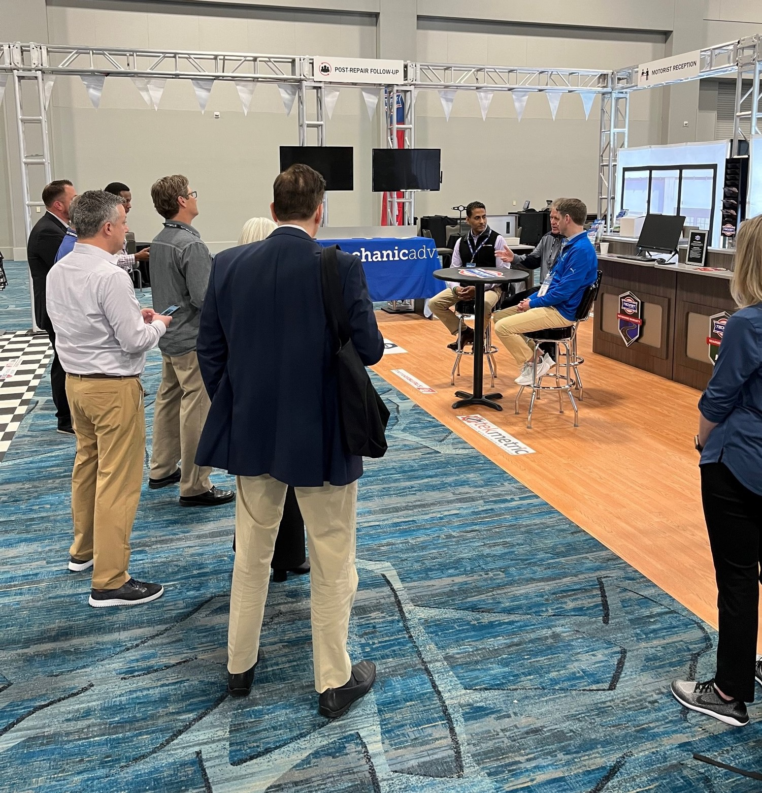 Seated at the table, Sunil Patel (left), with Tekmetric, and Parker Swift (right), with Mechanic Advisor, discuss business solutions at the Supplier and Training Expo (STX) with Bob Cushing, executive vice president of Advance Professional and president of Worldpac, with media and marketing representatives in attendance.