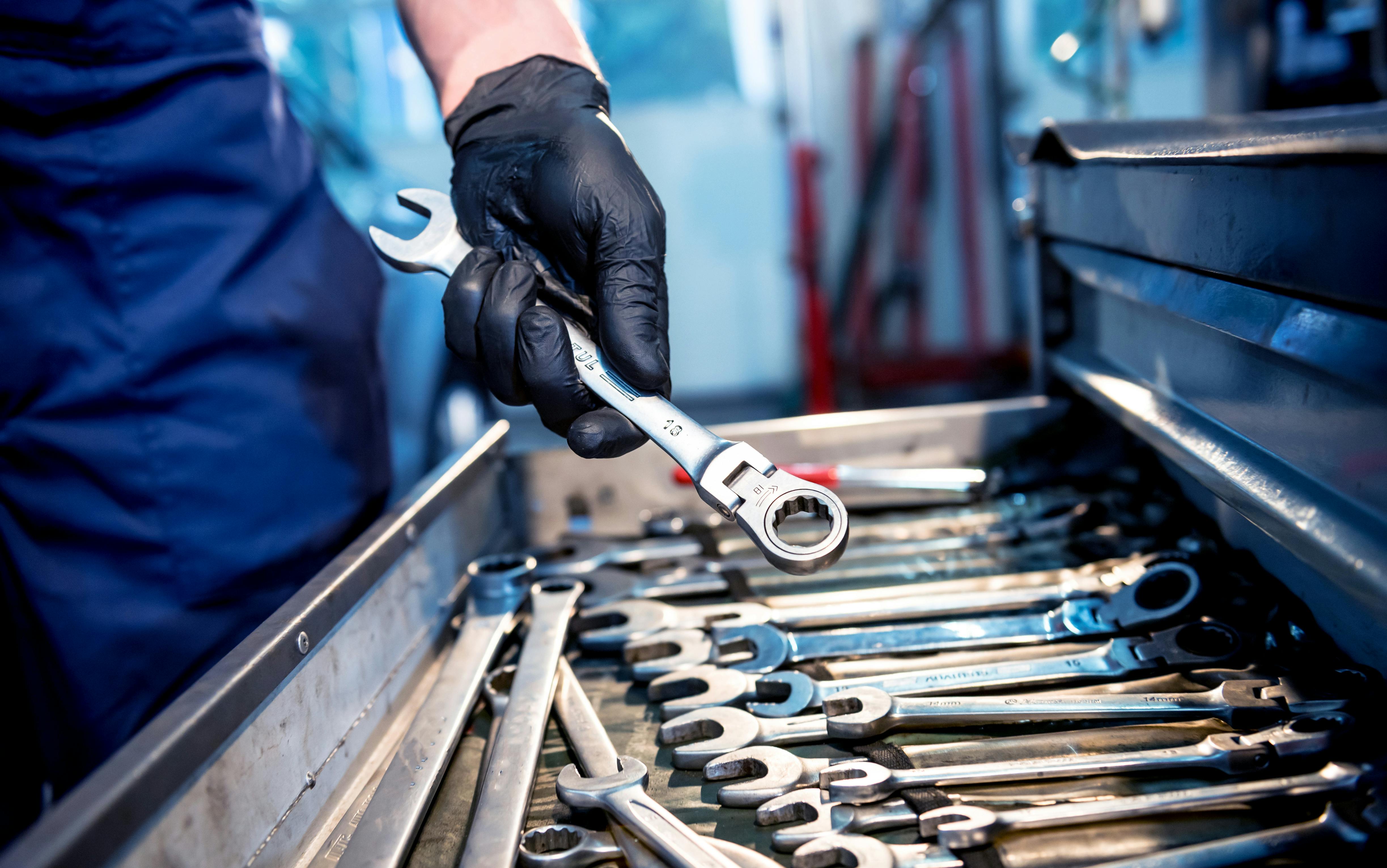 a person in mechanic's coveralls holds a ratcheting wrench above an open tool drawer of wrenches