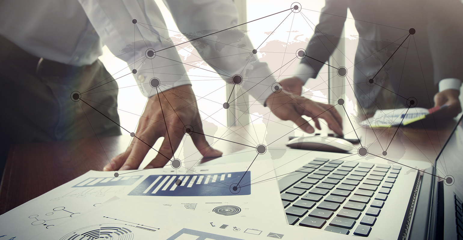 Business documents on office table with smart phone and digital tablet and stylus, and two colleagues discussing data in the background.