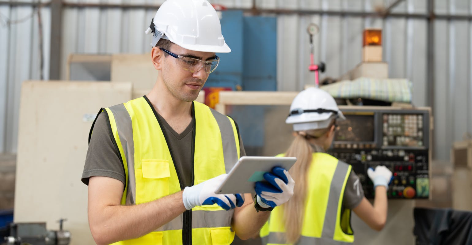 Engineer uses tablet while behind co-worker operates CNC machine.
