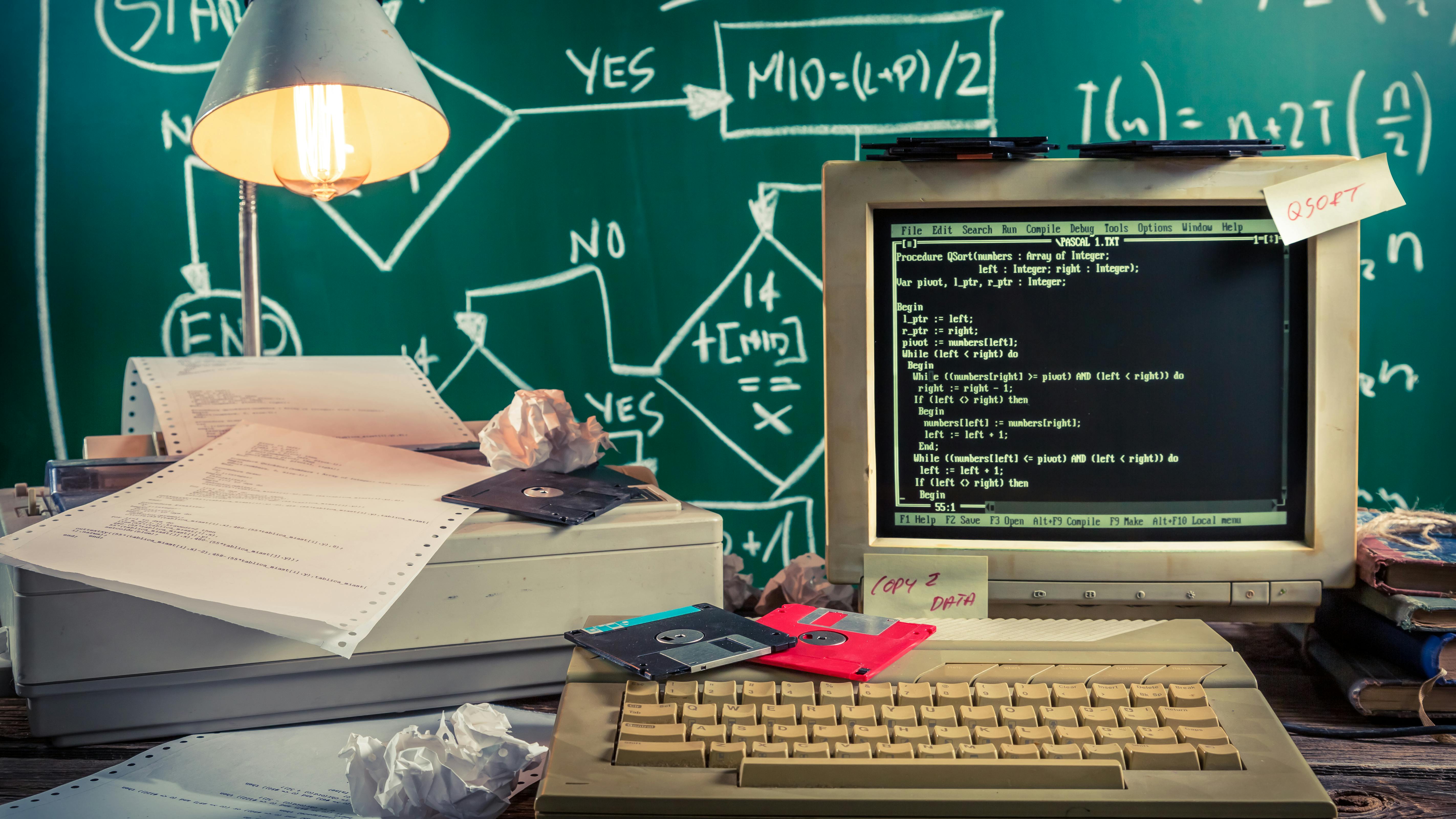an early personal computer with hard disks drives and dot matrix printer paper and equations written on a chalkboard in the background