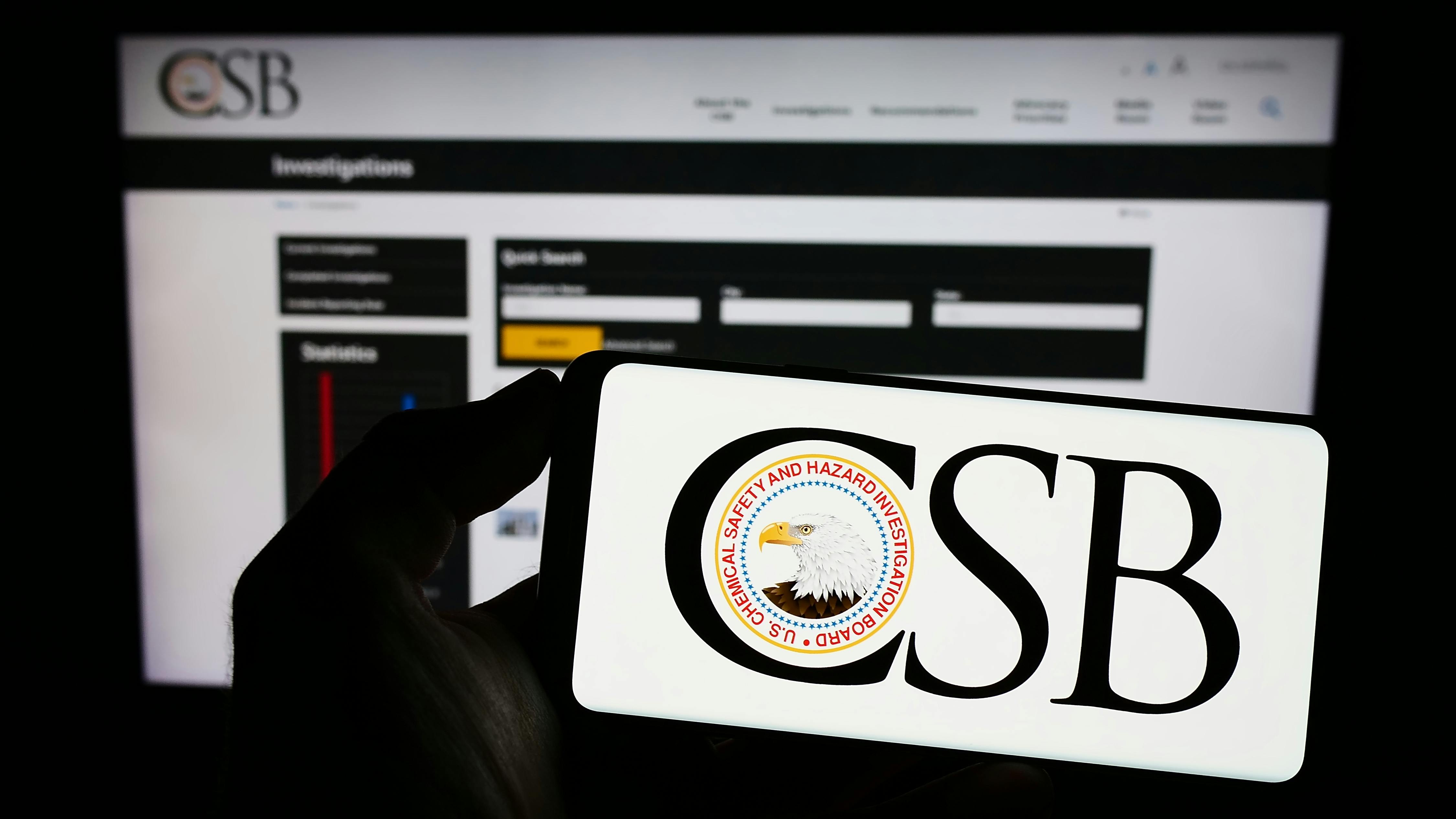 Person holding mobile phone with logo of federal agency U.S. CSB on screen in front of web page. F