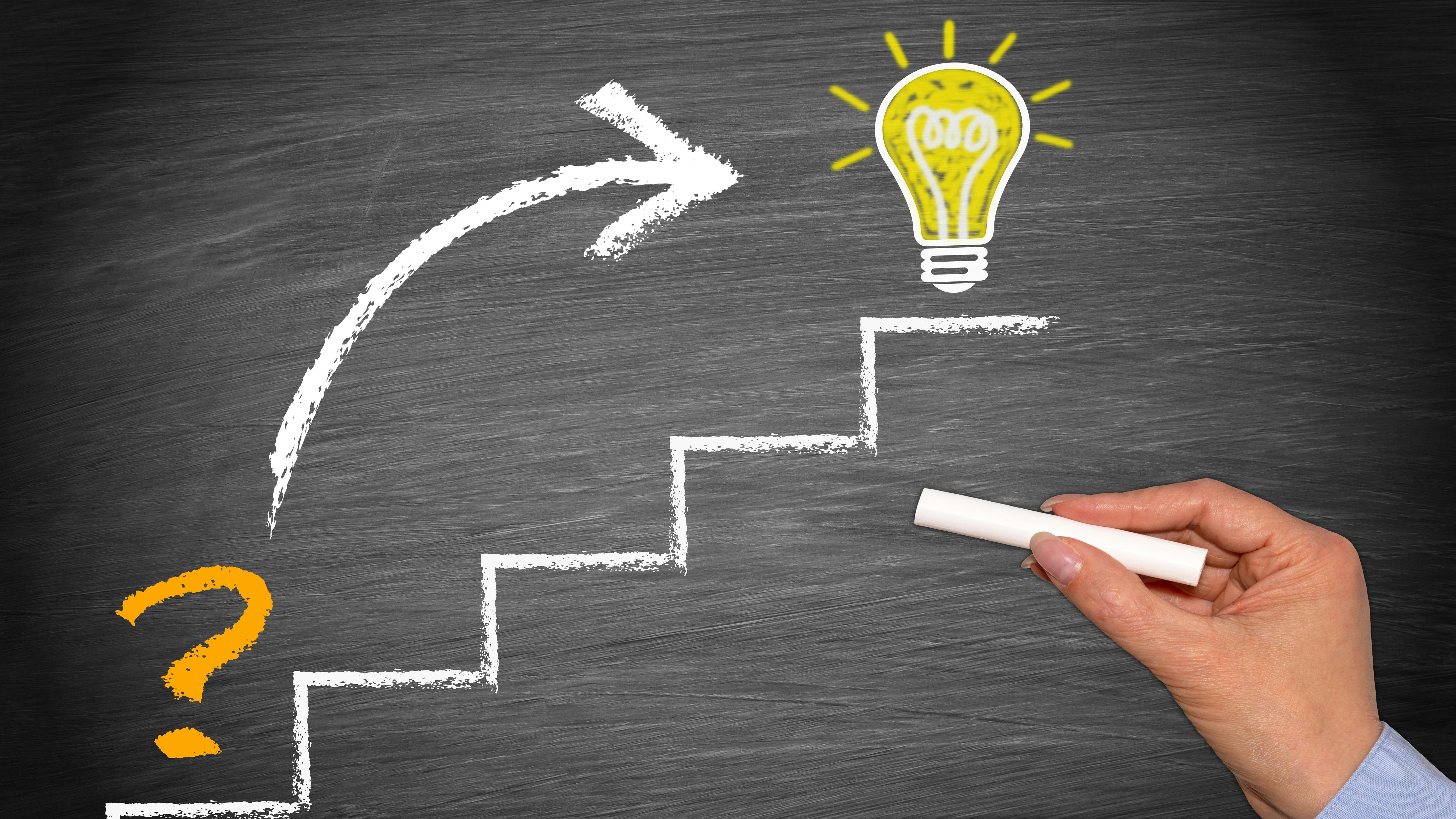 hand holds chalk in front of chalkboard with drawing of rudimentary stairs where bottom step has a question mark, an arrow points to top step that has an illuminated lightbulb