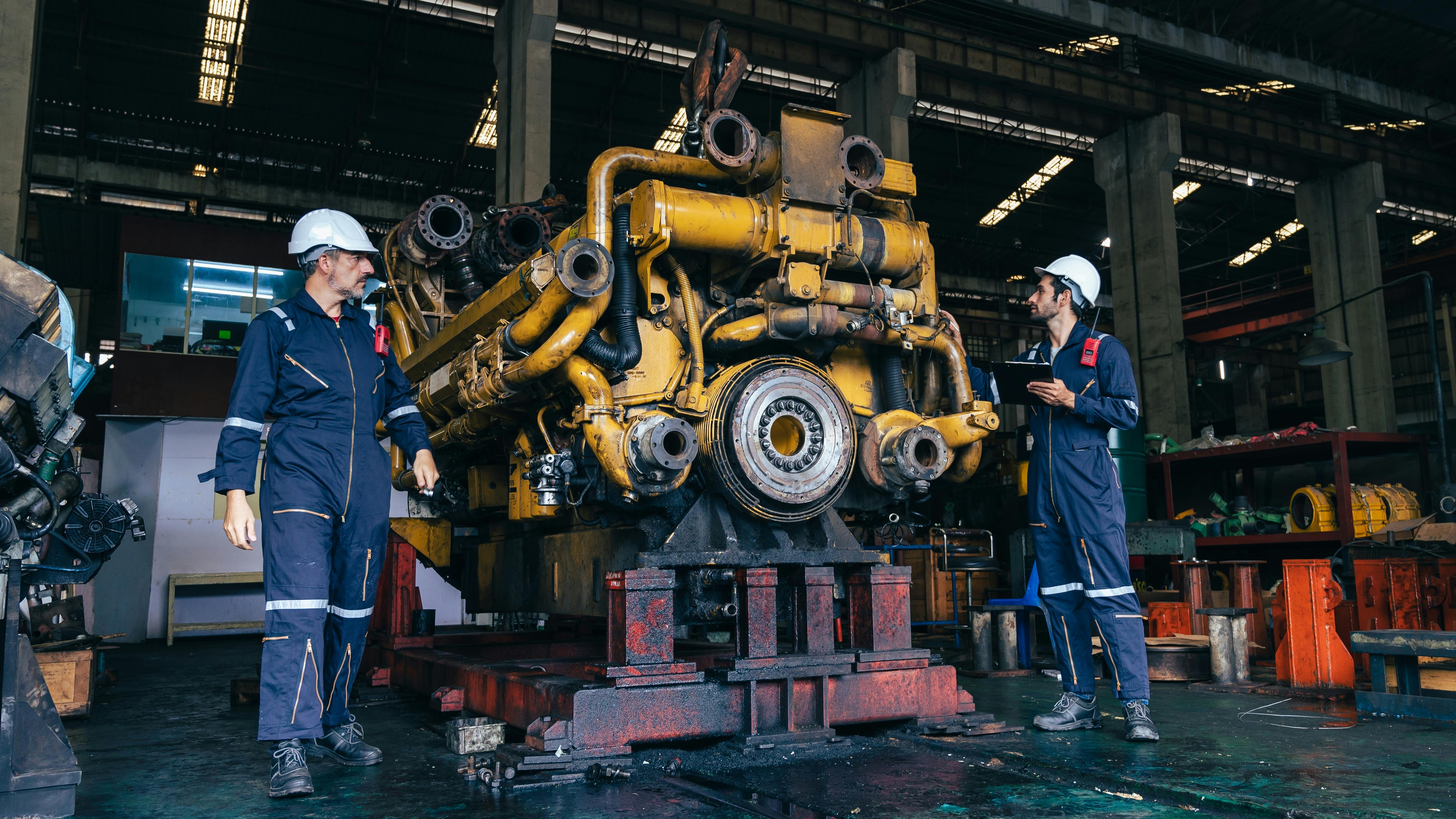2 men in work overalls and hard hats stand beside large diesel engine