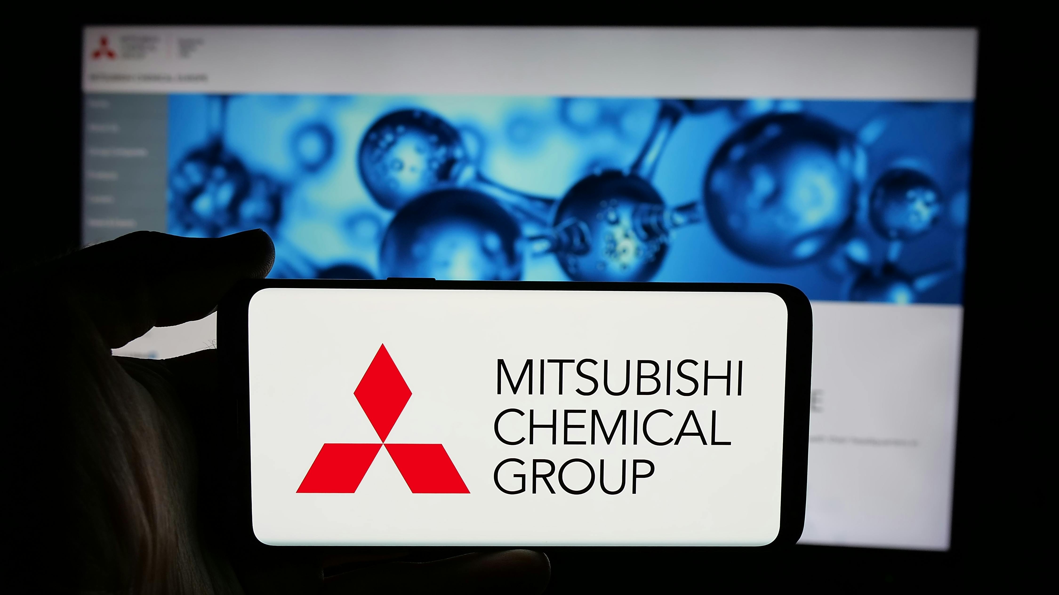 Mitsubishi Chemical Group logo displayed on phone screen with company website on computer screen in background