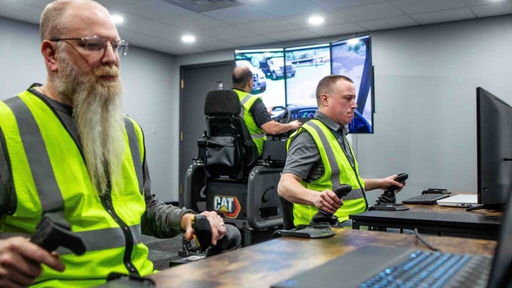 Three operator trainees work together in Cat Simulators Collaborative Worksite Training, cleaning up a spoil pile with a dozer and loading the material into an articulated truck with an excavator.