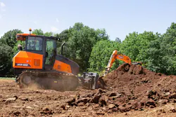 Develon DD100 dozer pushing dirt with an excavator in the background. Develon DD100 dozer pushing dirt with an excavator in the background.