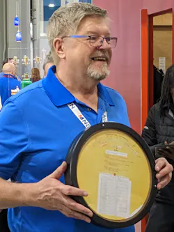 A smiling David Hughes holds his Carlson-Holohan Industry Award of Excellence—a replica of a Bell & Gossett System Syzer wheel (invented by Carlson). A smiling David Hughes holds his Carlson-Holohan Industry Award of Excellence—a replica of a Bell & Gossett System Syzer wheel (invented by Carlson).