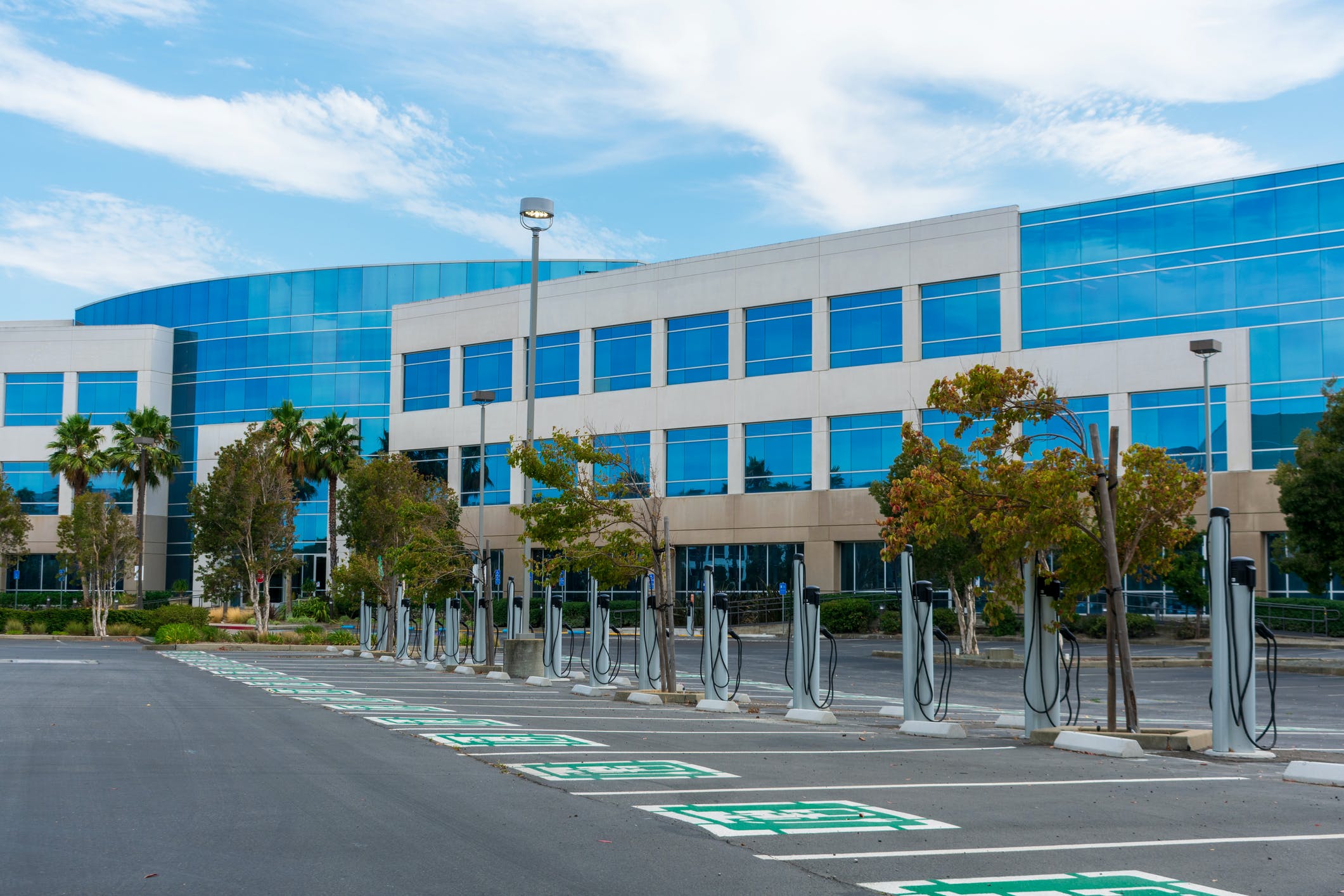 A row of electric vehicle charging stations with dedicated parking spots stands at outdoor parking lot of commercial office building.