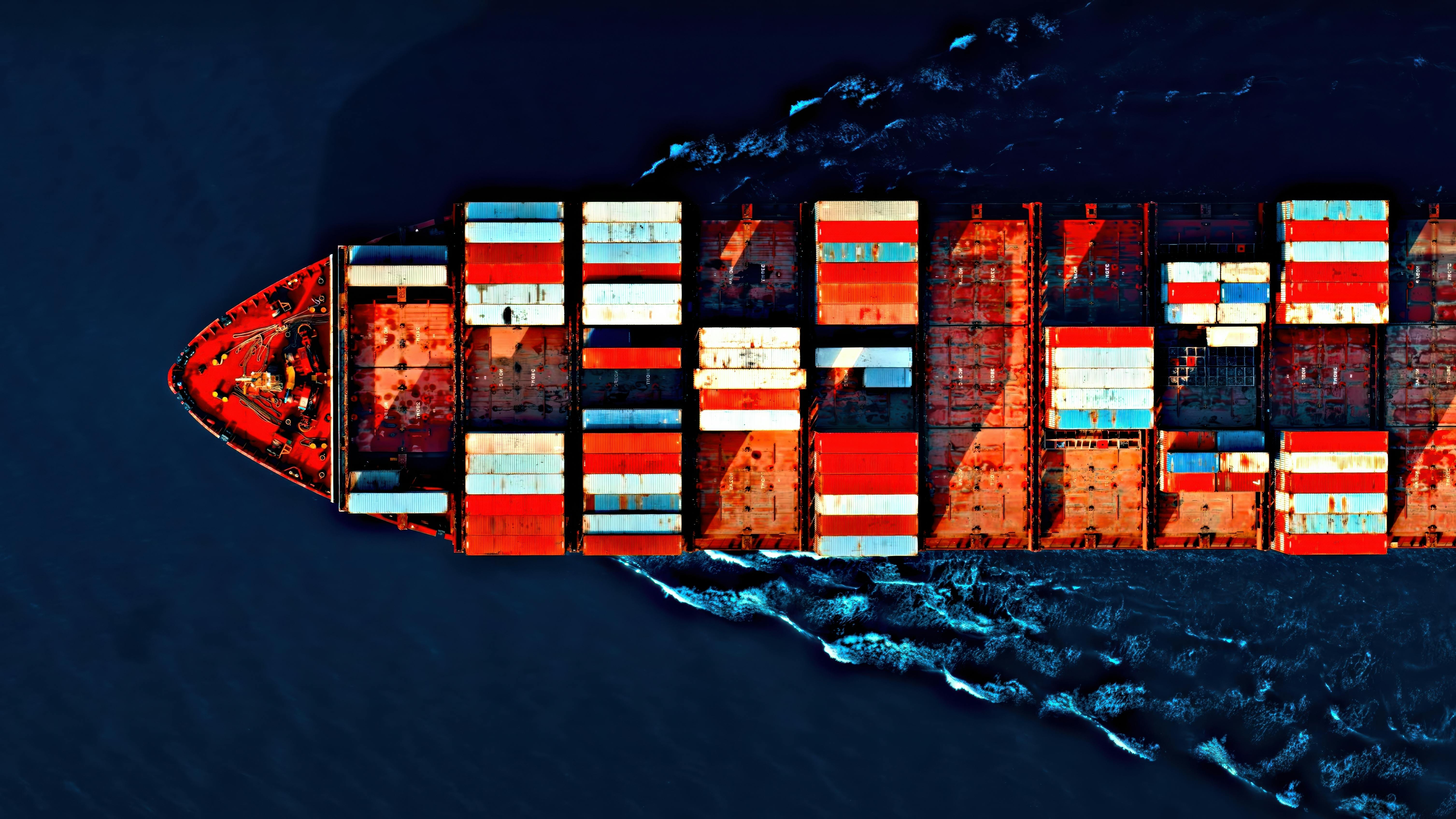 Aerial view of a cargo ship carrying colorful containers, cutting through deep blue waters with visible wave patterns.