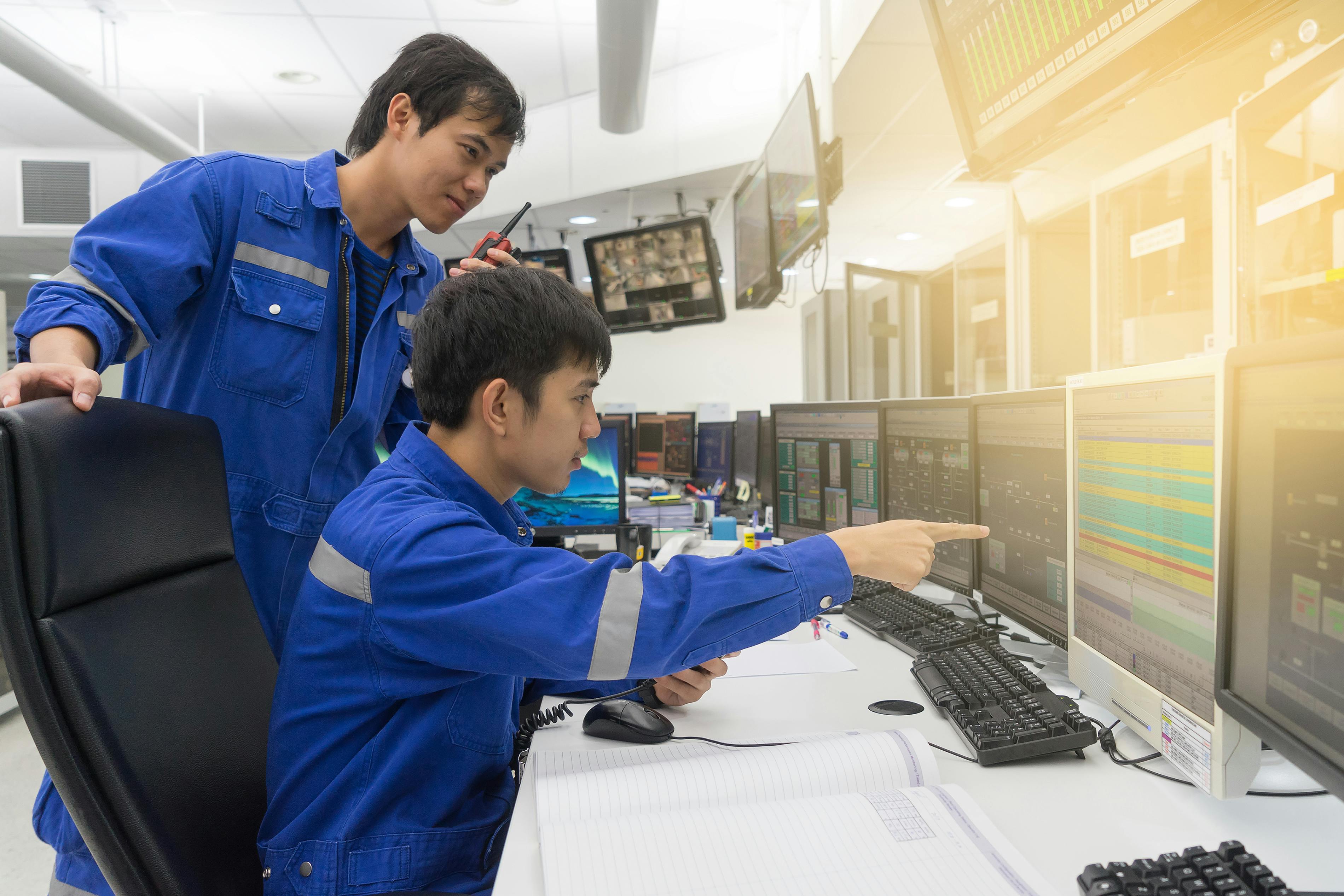 Two workers are examining machinery in an industrial facility. One person, wearing a white shirt and hard hat, operates a control panel, while the other observes in a bright safety vest.