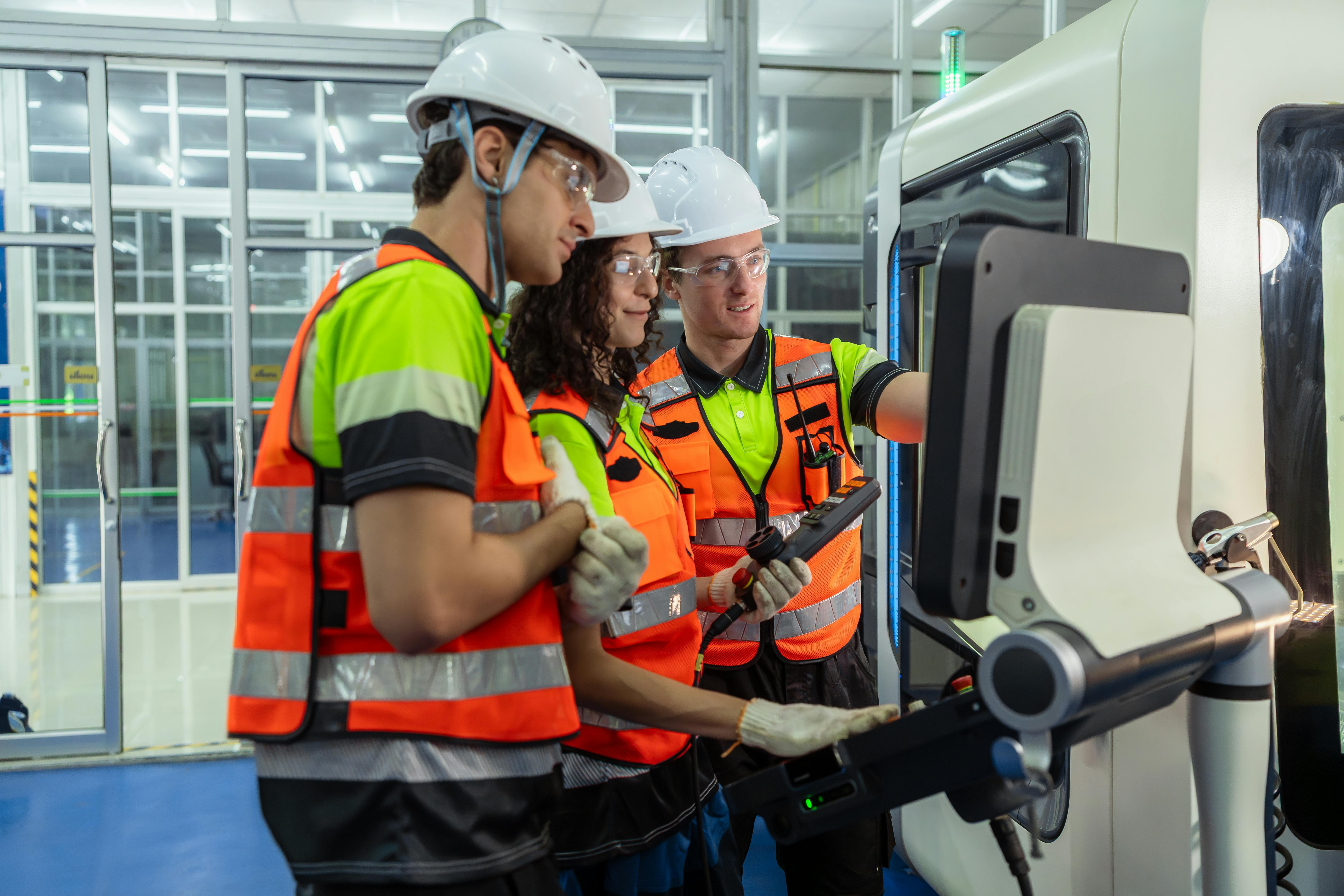 Three people wearing safety gear are standing in front of a computer monitor. One of the men is pointing at the screen