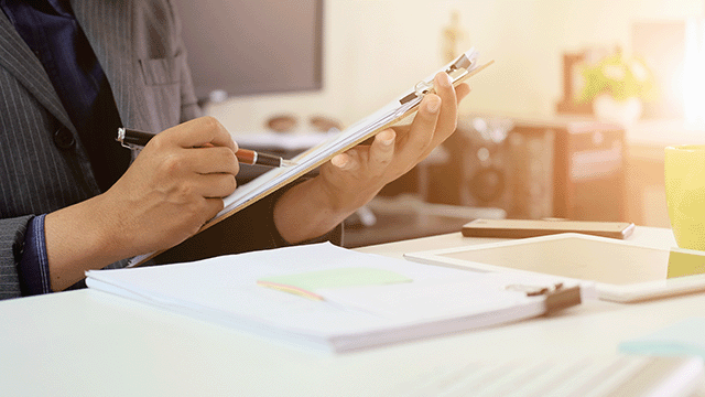 business person reviewing data on a clipboard with tablet and phone on table next to stack of papers
