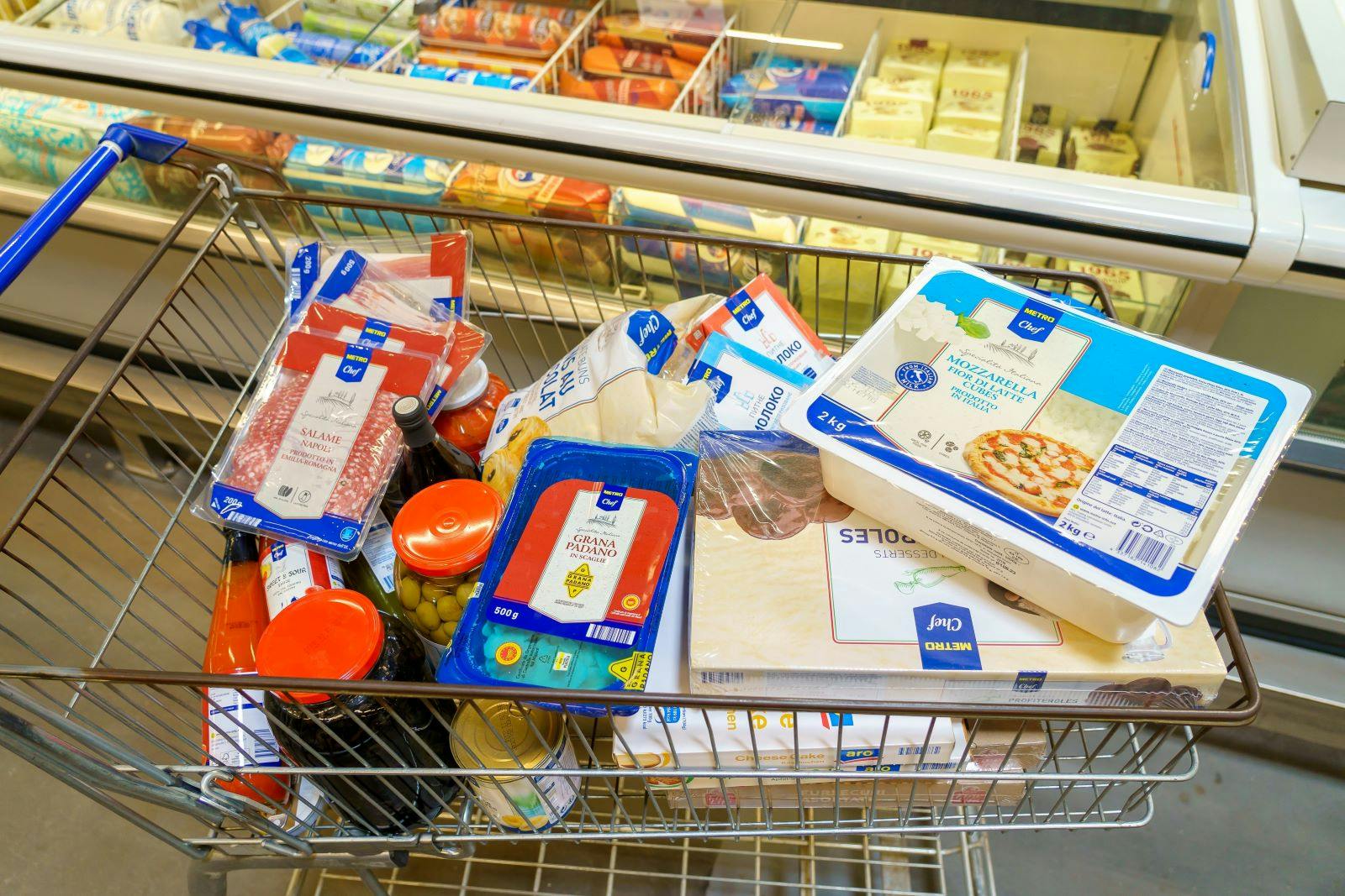 A shopping cart full of food items including pizza, cheese, and meat. The cart is in a store and the items are in the freezer section.