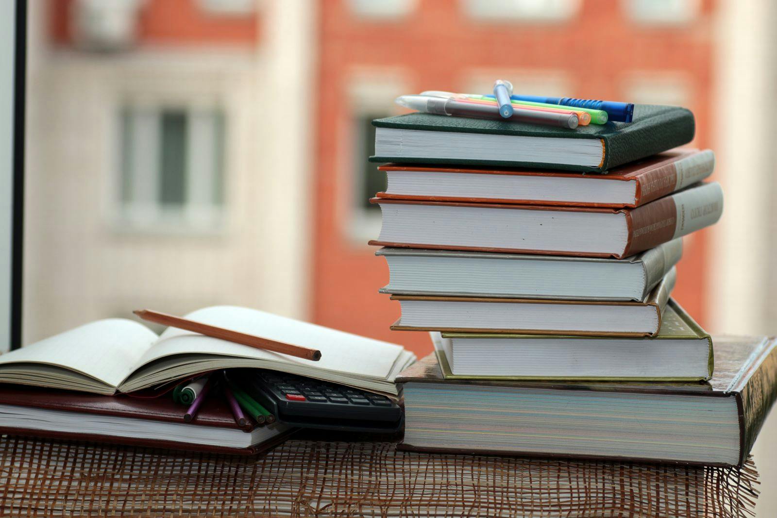 A stack of coursebooks and pens by a window