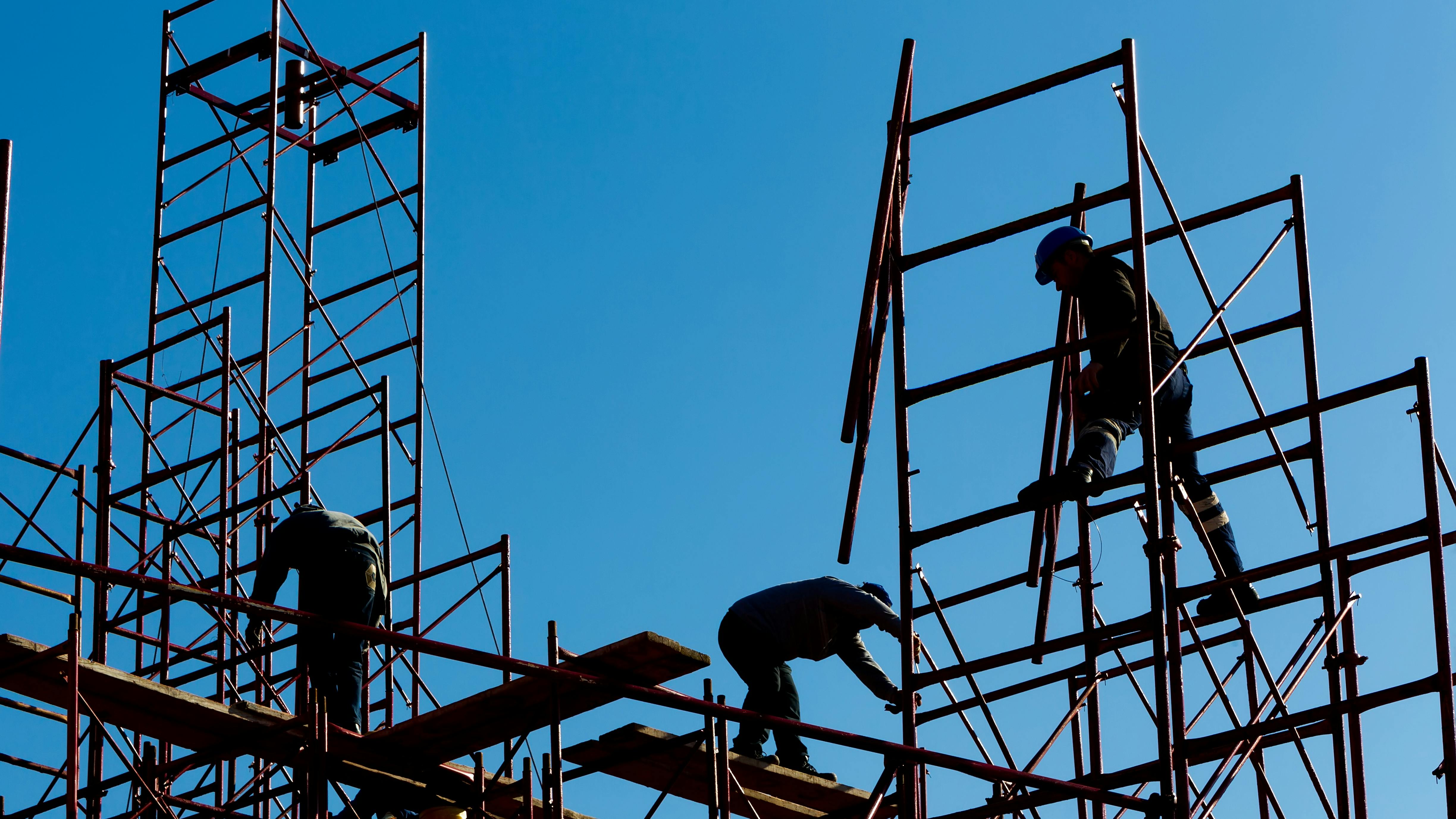 Silhouette of construction workers against sky on scaffolding with ladder on building site.