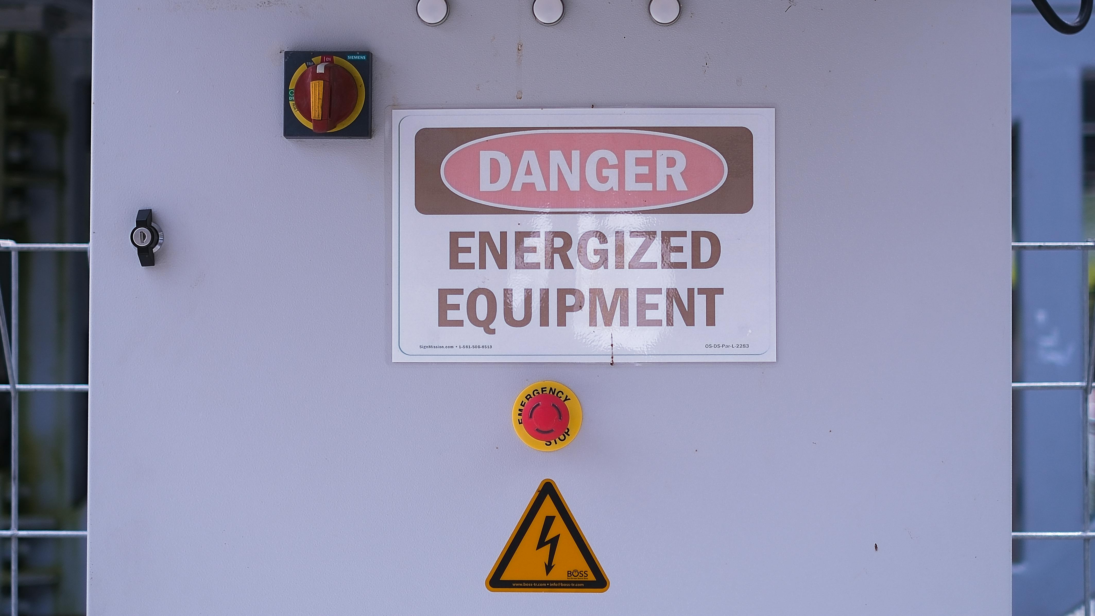 This image shows a close-up of an energized equipment warning sign, featuring a danger sign and emergency stop button. The safety measures are clearly visible, indicating high-voltage equipment. The image was generated by AI, highlighting the potential of AI in creating realistic visuals for safety