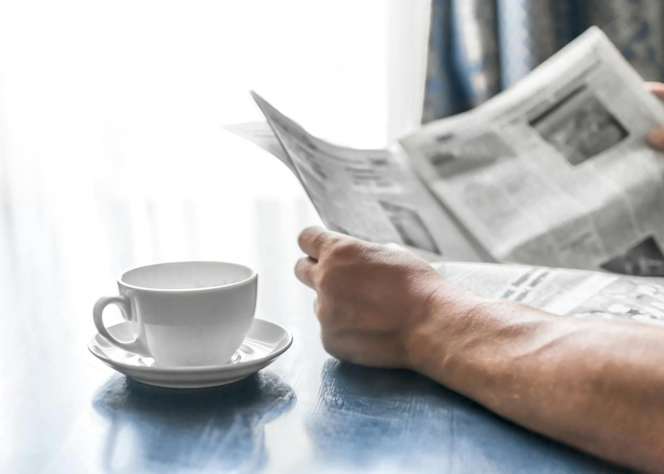 person reading newspaper near window while drinking a hot beverage