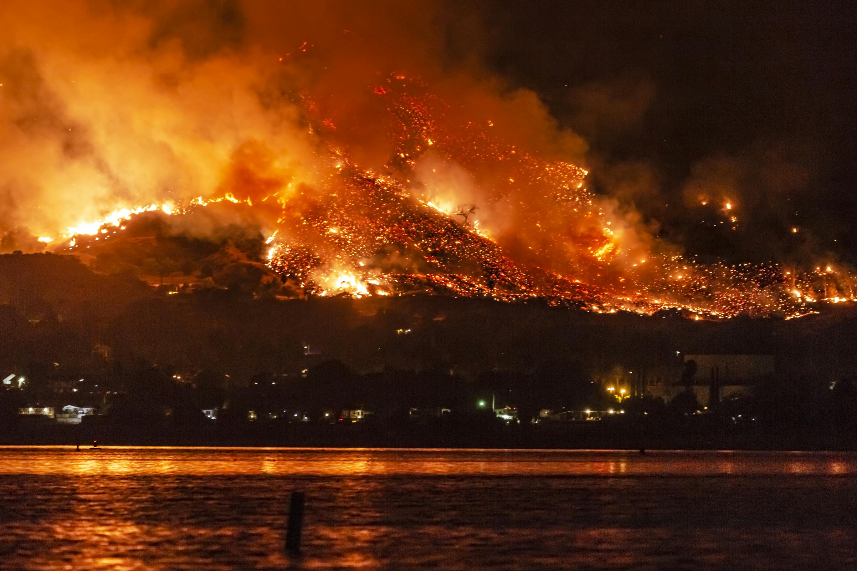 photo of a body of water in the forefront and a wildfire burning in the distance