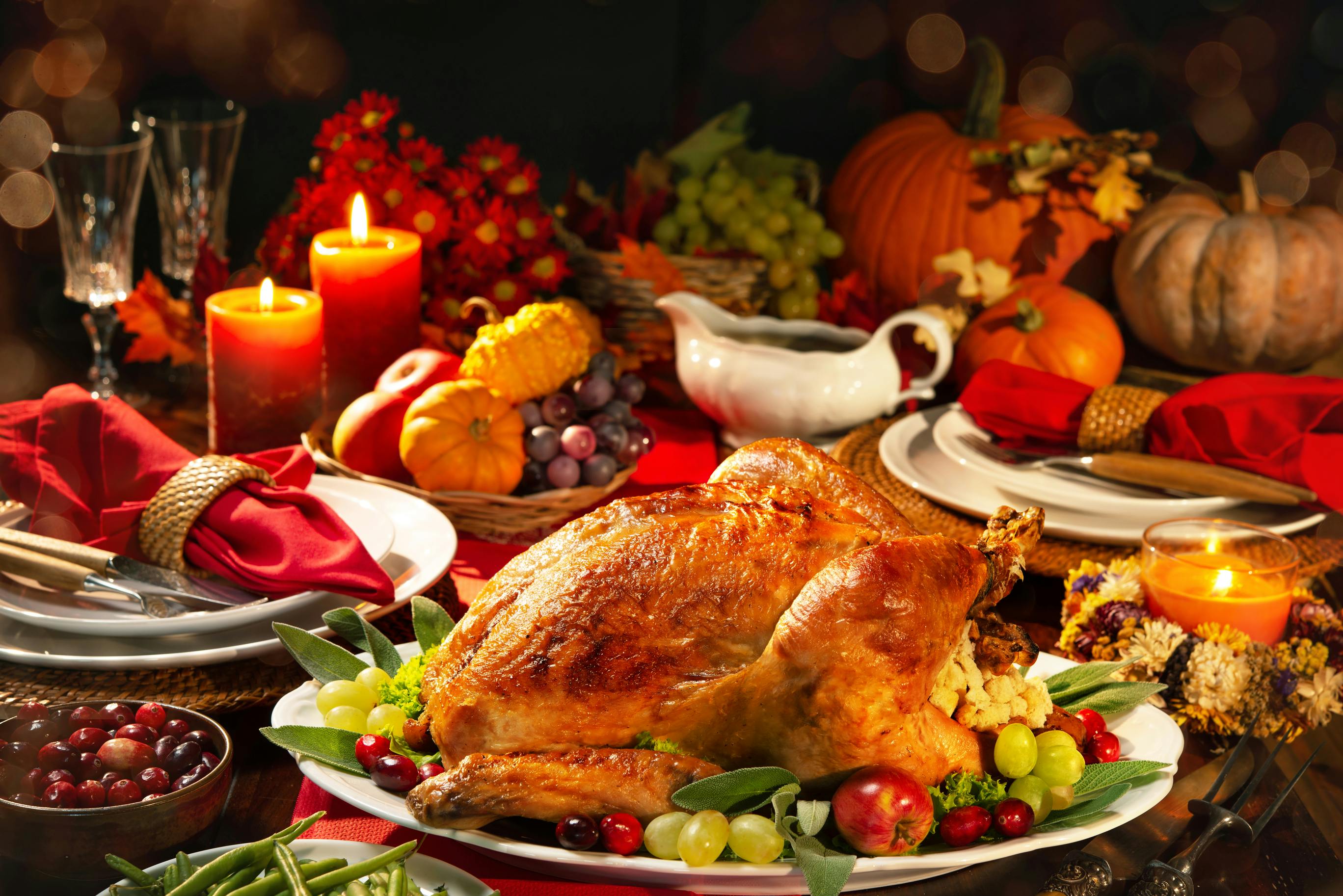 Picture of a tablescape with a turkey on a platter and formal place settings, gravy boat, candles, wine glasses and pumpkins in the background.