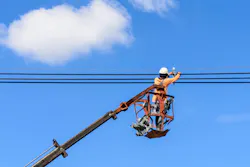 Field technician at height in bucket of hydraulic lift repairing electrical lines against a partly cloudy blue sky Field technician at height in bucket of hydraulic lift repairing electrical lines against a partly cloudy blue sky