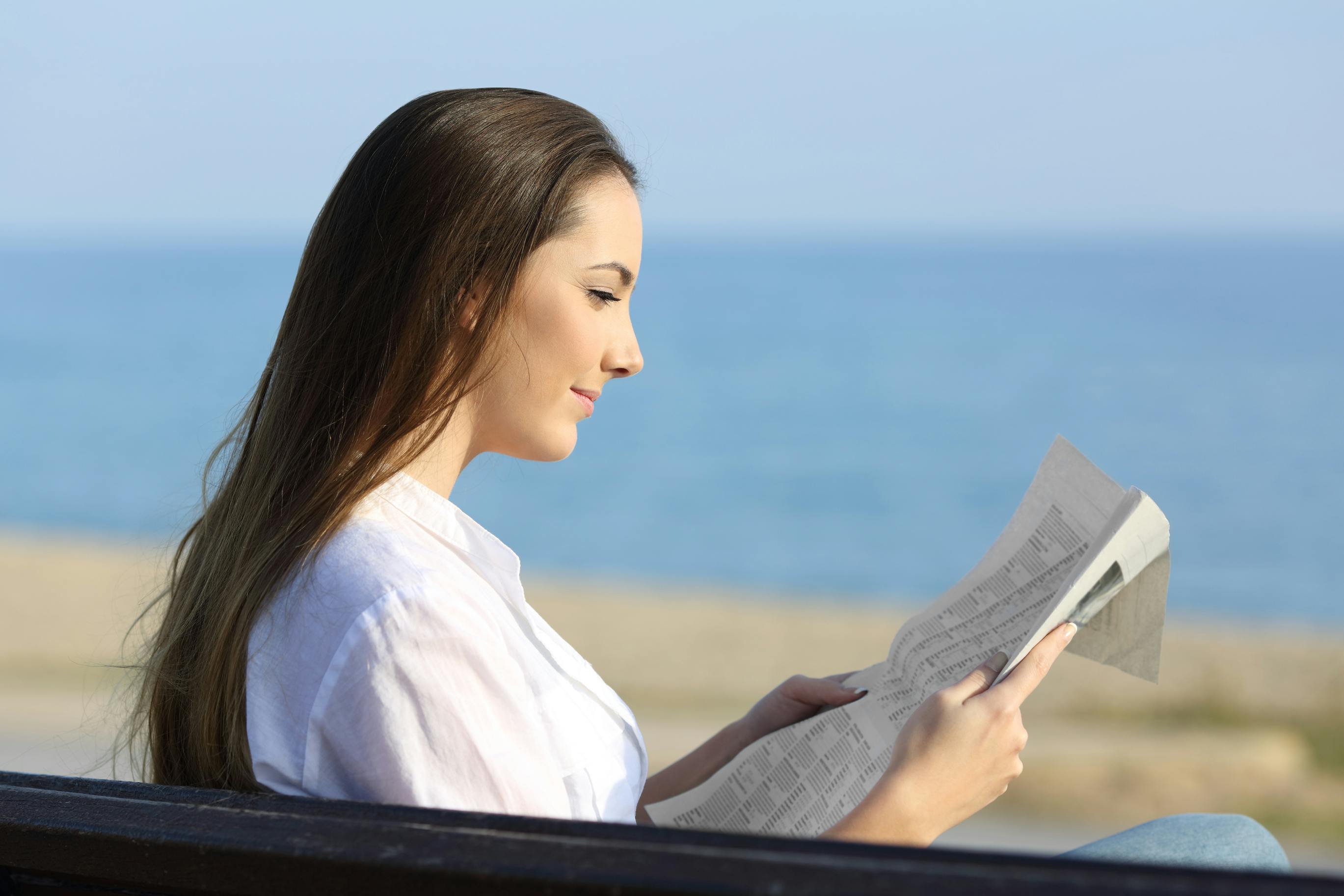 woman reading the newspaper outside on a bench near the beach