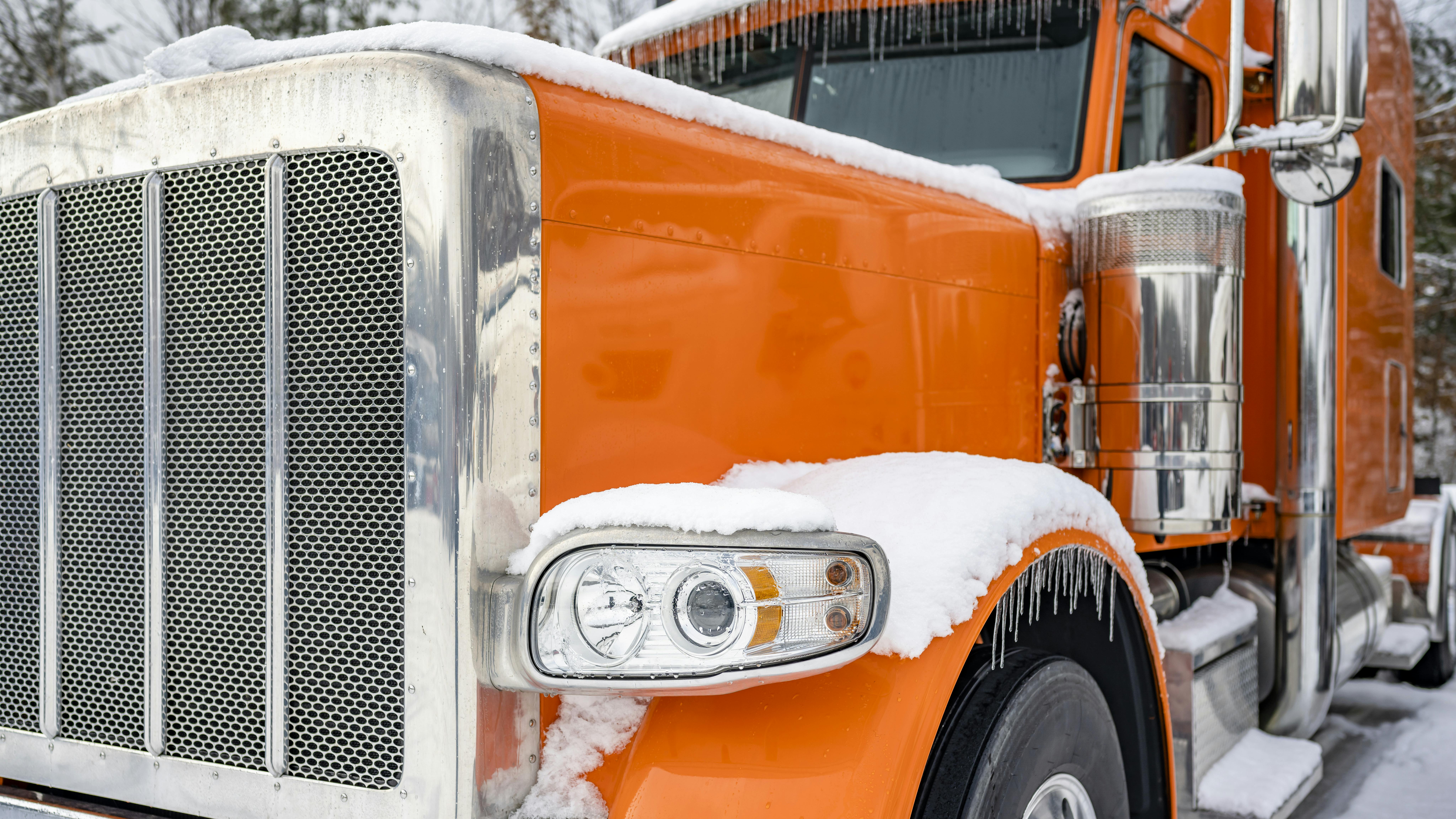 A classic orange big rig semi-truck with an extended cab parked in a snowy and icy truck stop parking lot during winter.
