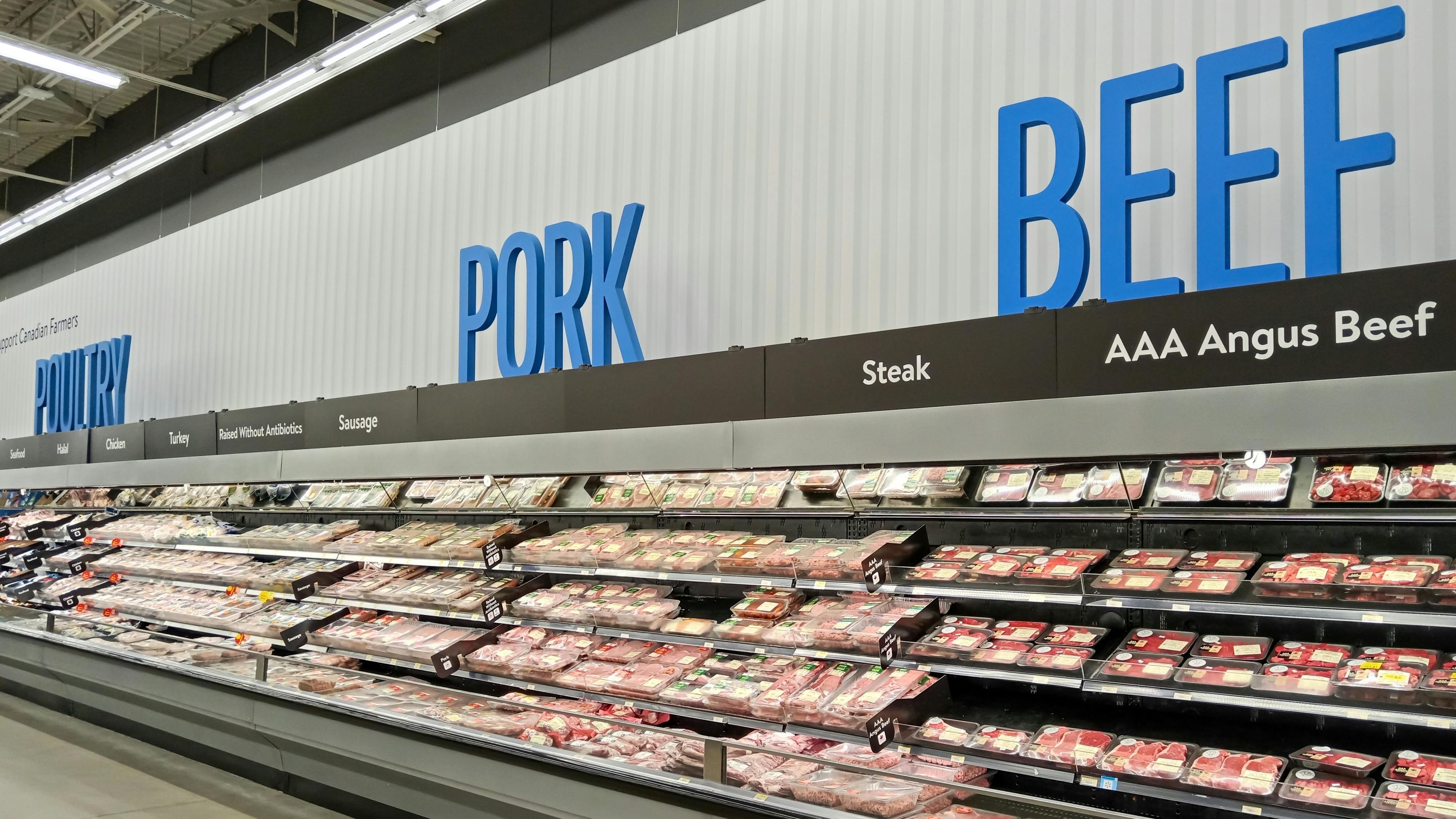 Grocery store meat department with long refrigerated display cases filled with packaged meat products. Large blue text on the white wall above reads 'POULTRY,' 'PORK,' and 'BEEF' from left to right.