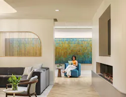 Residentially inspired lounge area in health facility with couches, fireplace, and woman reading in front of multicolored wall art. Residentially inspired lounge area in health facility with couches, fireplace, and woman reading in front of multicolored wall art.