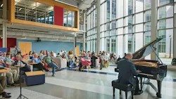 Man playing piano in front of audience in large, two-story space with glass wall in background. Man playing piano in front of audience in large, two-story space with glass wall in background.