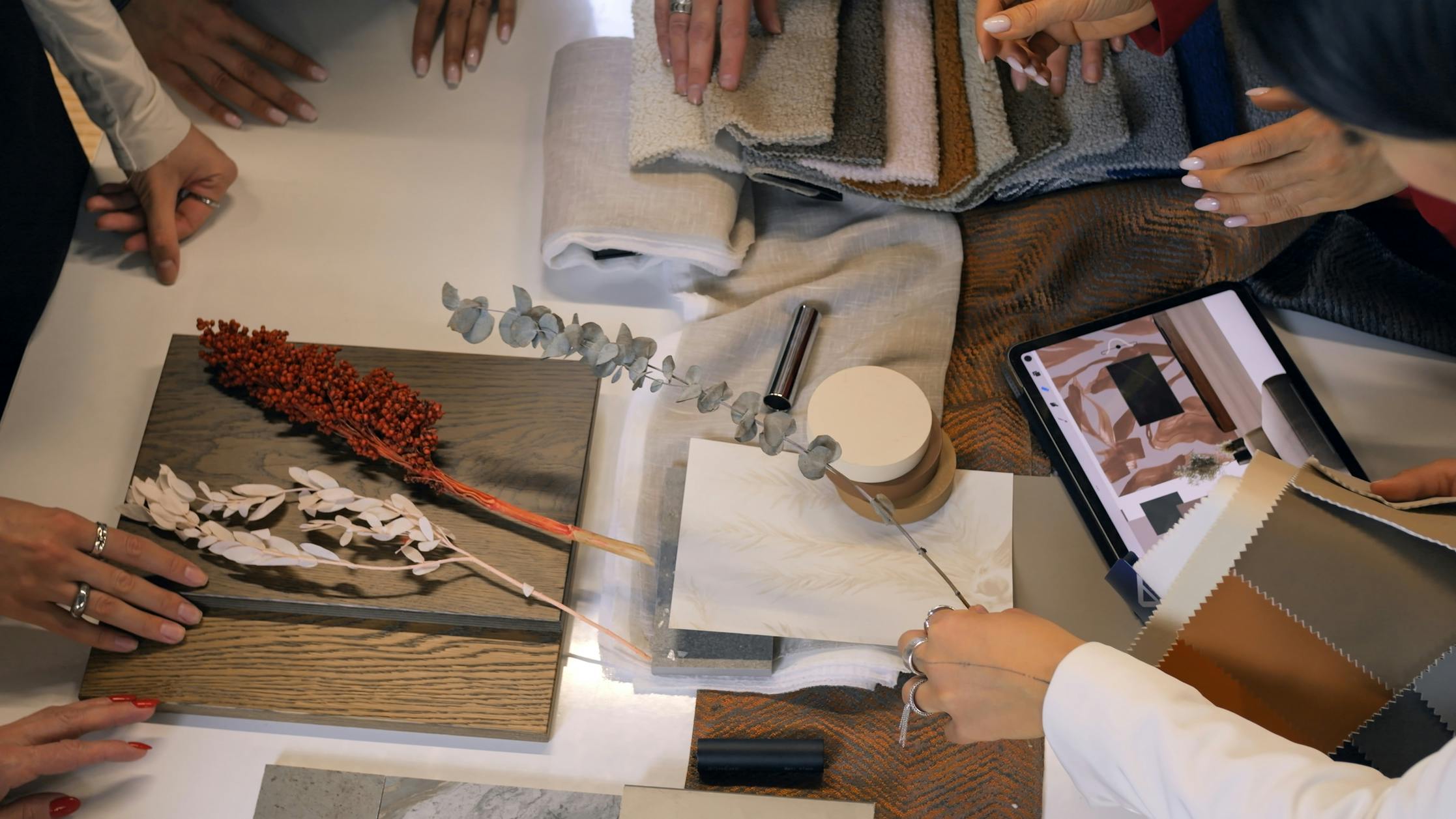 Interior designers are selecting materials and colors, using a tablet and various samples like wood, fabrics, and tiles, to create a cohesive design for their project.