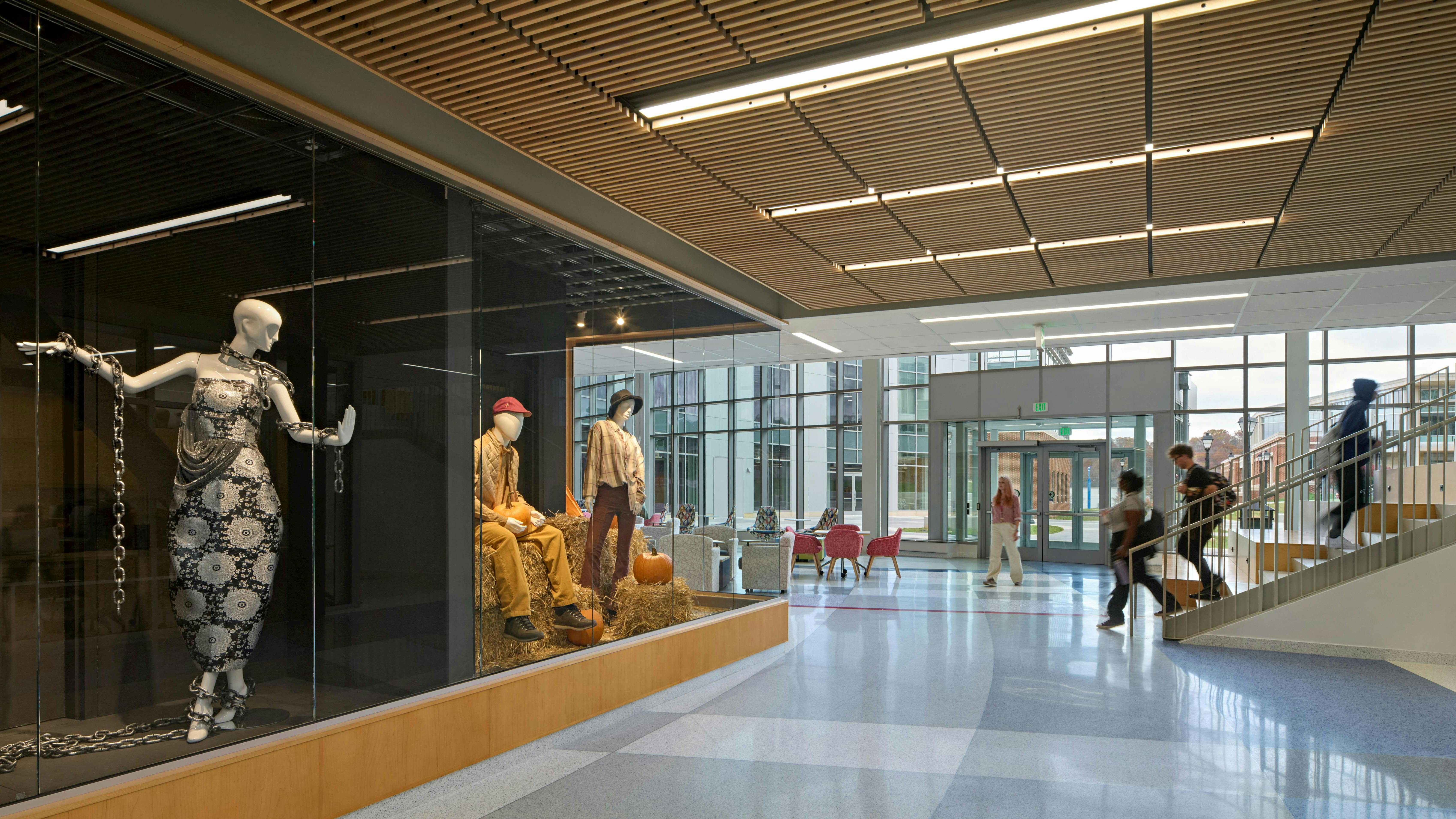 Hallway with glass wall showing fashion mannequins in foreground with staircase and glass wall with students walking in background.
